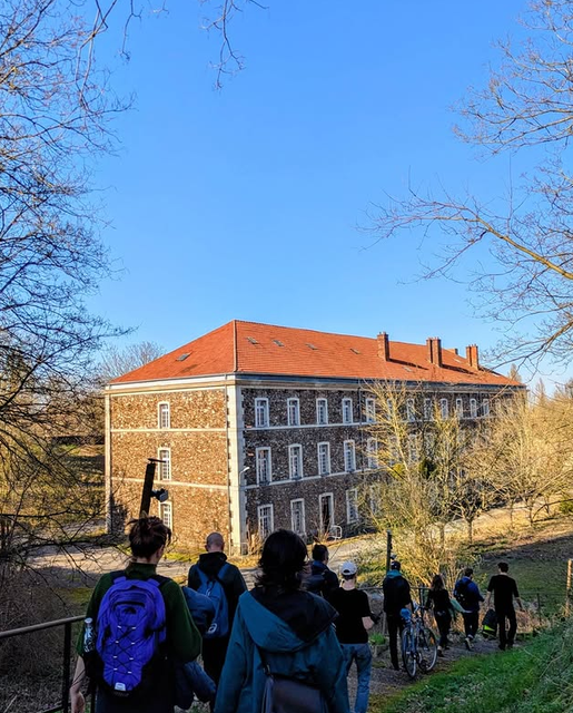 Plusieurs membres du collectif, de dos, marchent sur un chemin en descente vers le bâtiment de La Forte, une ancienne caserne militaire en brique.